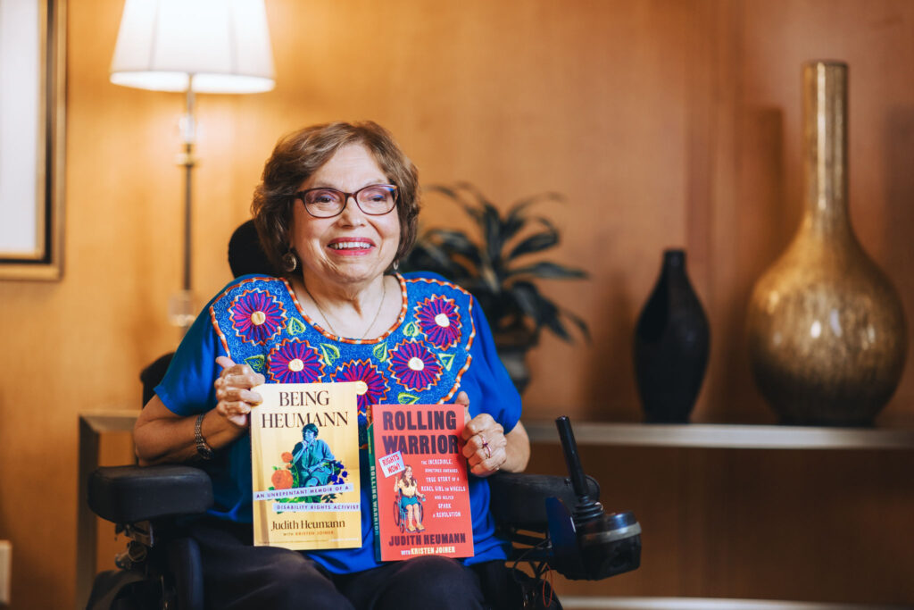 A photo of Judy Heumann holding her two books, Being Heumann and Rolling Warrior. Judy is a white woman with short brown hair who uses a wheelchair. She is wearing glasses, a bright blue shirt with pink embroidered flowers, and black pants. The book covers are yellow and red.

